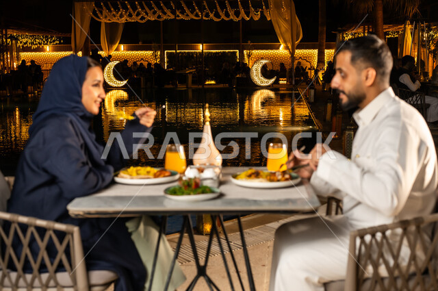 زينة وديكور شهر رمضان المبارك, قضاء وقت ممتع مع الأسرة, مائدة مليئة بالأطعمة والمشروبات الشهية، جلسة عائلية رمضانية روحانية, زوجان عربيان سعوديان خليجيان مبتسمان يرتديان الزي التقليدي يجلسان في مطعم, الاستمتاع بتناول الطعام, حوارات ونقاشات على مائدة الطعام