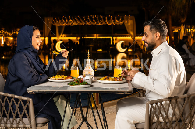 قضاء وقت ممتع مع الأسرة, مائدة مليئة بالأطعمة والمشروبات الشهية، جلسة عائلية رمضانية روحانية، زينة وديكور شهر رمضان المبارك,  زوجان عربيان سعوديان خليجيان مبتسمان يرتديان الزي التقليدي يجلسان في مطعم, الاستمتاع بتناول الطعام, حوارات ونقاشات على مائدة الطعام