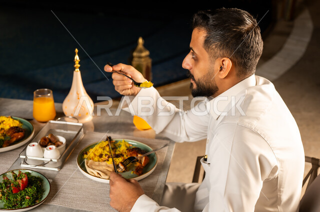 افطار رمضاني, مأكولات عربيه, وصفات شهيه في شهر رمضان المبارك, صورة من الجانب لرجل عربي خليجي سعودي يرتدي الزي التقليدي يقوم بتناول الطعام , مائده مليئه بأصناف المأكولات و المقبلات الشهية