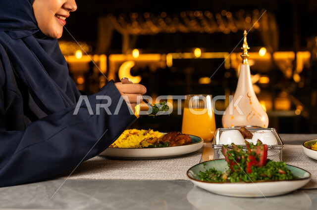 تقديم الطبق الرئيسي للافطار, أداء فريضة الصيام في شهر رمضان المبارك، سفرة مليئة بأصناف من المأكولات الشرقية الشهية، صورة من الجانب ليد امراة عربيه خليجيه سعودية ترتدي العباءة السوداء تقوم بتناول الطعام بايماءات السعاده و السرور, تناول المشروبات الطازجه