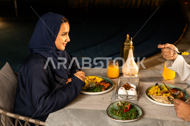 أداء فريضة الصيام في شهر رمضان المبارك، سفرة مليئة بأصناف من المأكولات الشرقية الشهية، صورة من الجانب لامراة عربيه خليجيه سعودية ترتدي العباءة السوداء تقوم بتناول الطعام بايماءات السعاده و السرور, الاستمتاع بالمشروبات الطازجه