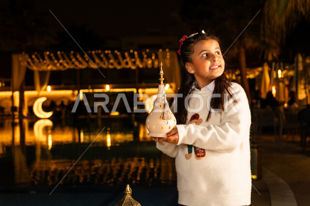 Decorations and lights for holidays and occasions, preparing and getting ready to welcome the holy month of Ramadan, celebrating the arrival of the month of goodness, a smiling Saudi Arabian Gulf girl wearing casual clothes holding a lit, decorated lantern in her hand and looking at something, expressions of joy and happiness