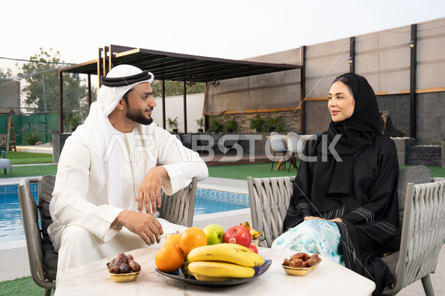 قضاء أوقات عائلية ممتعة, التفاهم والترابط الفكري بين الزوجين، تناول الوجبات الخفيفة والتسالي، الجلوس في إحدى الشاليهات الاماراتية الراقية، رجل عربي خليجي اماراتي يرتدي الكندورة والغترة يقوم بتبادل الأحاديث مع زوجته في الهواء الطلق