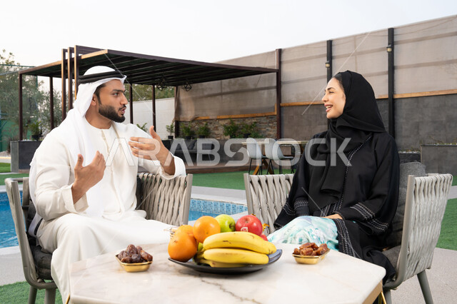 An Emirati Gulf Arab man wearing a kandura and ghutra exchanging conversations with his wife outdoors, spending enjoyable family time, understanding and intellectual connection between the spouses, eating snacks and entertainment, sitting in one of the upscale Emirati chalets