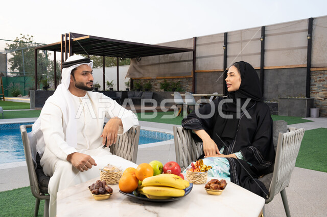 قضاء أوقات عائلية ممتعة, التفاهم والترابط الفكري بين الزوجين، تناول الوجبات الخفيفة والتسالي، الجلوس في إحدى الشاليهات الاماراتية الراقية، رجل عربي خليجي اماراتي يرتدي الكندورة والغترة يقوم بتبادل الأحاديث مع زوجته في الهواء الطلق
