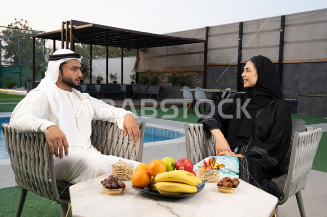 قضاء أوقات عائلية ممتعة, التفاهم والترابط الفكري بين الزوجين، تناول الوجبات الخفيفة والتسالي، الجلوس في إحدى الشاليهات الاماراتية الراقية، رجل عربي خليجي اماراتي يرتدي الكندورة والغترة يقوم بتبادل الأحاديث مع زوجته في الهواء الطلق