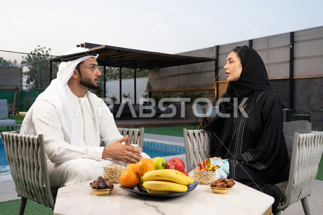 قضاء أوقات عائلية ممتعة, التفاهم والترابط الفكري بين الزوجين، تناول الوجبات الخفيفة والتسالي، الجلوس في إحدى الشاليهات الاماراتية الراقية، رجل عربي خليجي اماراتي يرتدي الكندورة والغترة يقوم بتبادل الأحاديث مع زوجته في الهواء الطلق