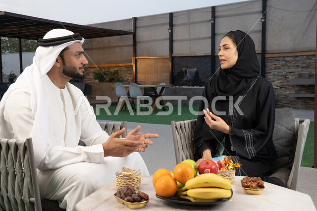 رجل عربي خليجي اماراتي يرتدي الكندورة والغترة يقوم بتبادل الأحاديث مع زوجته في الهواء الطلق, قضاء أوقات عائلية ممتعة, التفاهم والترابط الفكري بين الزوجين، تنال الوجبات الخفيفة والتسالي، الجلوس في إحدى الشاليهات الاماراتية الراقية