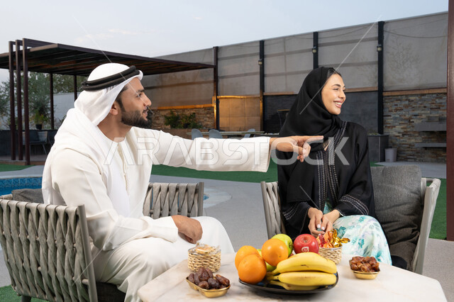 الاشارة الى شيء ما، الجلوس في إحدى الشاليهات الاماراتية الراقية, رجل عربي خليجي اماراتي يرتدي الكندورة والغترة يقوم بتبادل الأحاديث مع زوجته في الهواء الطلق, قضاء أوقات عائلية ممتعة, التفاهم والترابط الفكري بين الزوجين، تناول الوجبات الخفيفة والتسالي