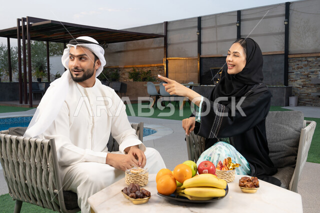 تعابير السرور والسعادة، الاشارة الى شيء ما، الجلوس في إحدى الشاليهات الاماراتية الراقية, رجل عربي خليجي اماراتي يرتدي الكندورة والغترة يقوم بتبادل الأحاديث مع زوجته في الهواء الطلق, قضاء أوقات عائلية ممتعة، تناول الوجبات الخفيفة والتسالي