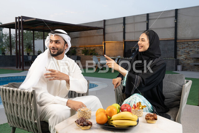 تعابير السرور والسعادة، الاشارة الى شيء ما، الجلوس في إحدى الشاليهات الاماراتية الراقية, رجل عربي خليجي اماراتي يرتدي الكندورة والغترة يقوم بتبادل الأحاديث مع زوجته في الهواء الطلق, قضاء أوقات عائلية ممتعة، تناول الوجبات الخفيفة والتسالي