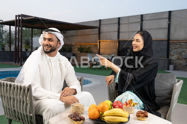 الاشارة الى شيء ما، الجلوس في إحدى الشاليهات الاماراتية الراقية, رجل عربي خليجي اماراتي يرتدي الكندورة والغترة يقوم بتبادل الأحاديث مع زوجته في الهواء الطلق, قضاء أوقات عائلية ممتعة, التفاهم والترابط الفكري بين الزوجين، تناول الوجبات الخفيفة والتسالي
