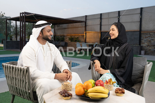 قضاء أوقات عائلية ممتعة, التفاهم والترابط الفكري بين الزوجين، تناول الوجبات الخفيفة والتسالي، الجلوس في إحدى الشاليهات الاماراتية الراقية، رجل عربي خليجي اماراتي يرتدي الكندورة والغترة يقوم بتبادل الأحاديث مع زوجته في الهواء الطلق