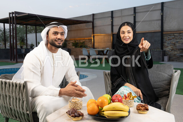 تعابير السرور والسعادة، الاشارة الى شيء ما، الجلوس في إحدى الشاليهات الاماراتية الراقية, رجل عربي خليجي اماراتي يرتدي الكندورة والغترة يقوم بتبادل الأحاديث مع زوجته في الهواء الطلق, قضاء أوقات عائلية ممتعة، تناول الوجبات الخفيفة والتسالي