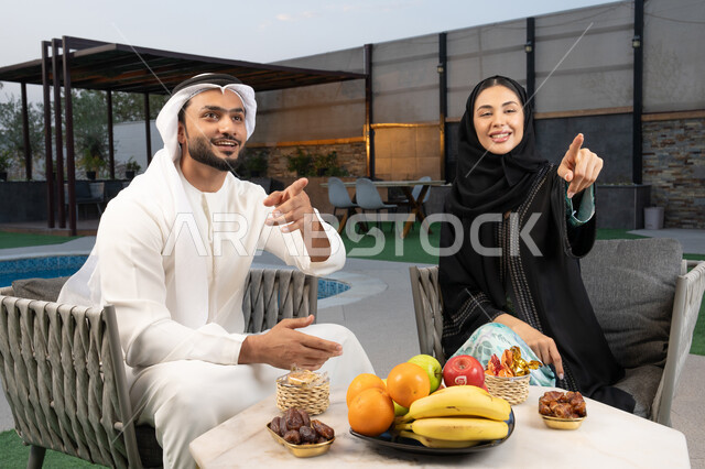 الاشارة الى شيء ما، الجلوس في إحدى الشاليهات الاماراتية الراقية, رجل عربي خليجي اماراتي يرتدي الكندورة والغترة يقوم بتبادل الأحاديث مع زوجته في الهواء الطلق, قضاء أوقات عائلية ممتعة, التفاهم والترابط الفكري بين الزوجين، تناول الوجبات الخفيفة والتسالي