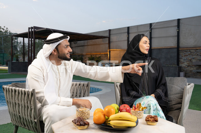 تعابير السرور والسعادة، الاشارة الى شيء ما، الجلوس في إحدى الشاليهات الاماراتية الراقية, رجل عربي خليجي اماراتي يرتدي الكندورة والغترة يقوم بتبادل الأحاديث مع زوجته في الهواء الطلق, قضاء أوقات عائلية ممتعة، تناول الوجبات الخفيفة والتسالي