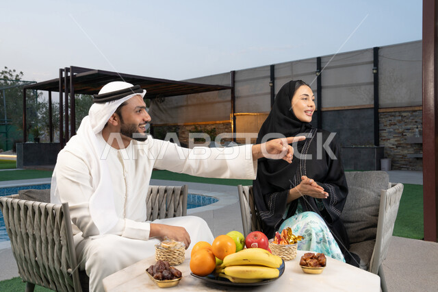 تعابير السرور والسعادة، الاشارة الى شيء ما، الجلوس في إحدى الشاليهات الاماراتية الراقية, رجل عربي خليجي اماراتي يرتدي الكندورة والغترة يقوم بتبادل الأحاديث مع زوجته في الهواء الطلق, قضاء أوقات عائلية ممتعة، تناول الوجبات الخفيفة والتسالي