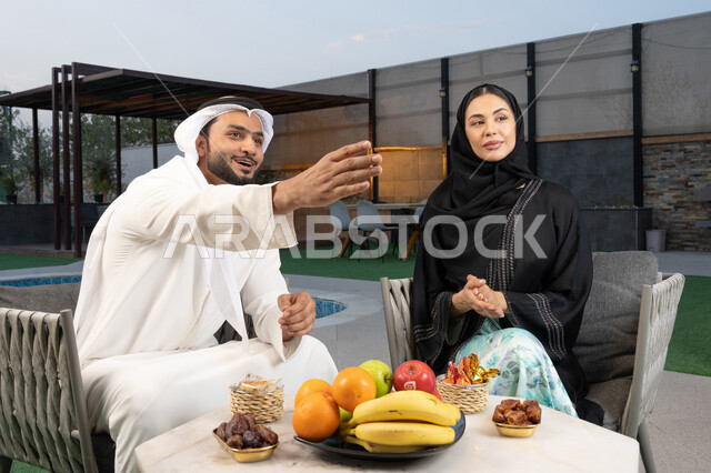 الاشارة الى شيء ما، الجلوس في إحدى الشاليهات الاماراتية الراقية, رجل عربي خليجي اماراتي يرتدي الكندورة والغترة يقوم بتبادل الأحاديث مع زوجته في الهواء الطلق, قضاء أوقات عائلية ممتعة, التفاهم والترابط الفكري بين الزوجين، تناول الوجبات الخفيفة والتسالي