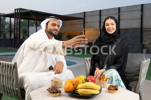 تعابير السرور والسعادة، الاشارة الى شيء ما، الجلوس في إحدى الشاليهات الاماراتية الراقية, رجل عربي خليجي اماراتي يرتدي الكندورة والغترة يقوم بتبادل الأحاديث مع زوجته في الهواء الطلق, قضاء أوقات عائلية ممتعة، تناول الوجبات الخفيفة والتسالي
