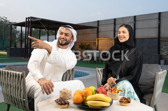 الاشارة الى شيء ما، الجلوس في إحدى الشاليهات الاماراتية الراقية, رجل عربي خليجي اماراتي يرتدي الكندورة والغترة يقوم بتبادل الأحاديث مع زوجته في الهواء الطلق, قضاء أوقات عائلية ممتعة, التفاهم والترابط الفكري بين الزوجين، تناول الوجبات الخفيفة والتسالي