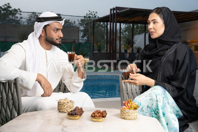 شرب القهوة العربية، قضاء أوقات عائلية ممتعة, التفاهم والترابط الفكري بين الزوجين، تناول الوجبات الخفيفة والتسالي، الجلوس في إحدى الشاليهات الاماراتية الراقية، رجل عربي خليجي اماراتي يرتدي الكندورة والغترة يقوم بتبادل الأحاديث مع زوجته في الهواء الطلق