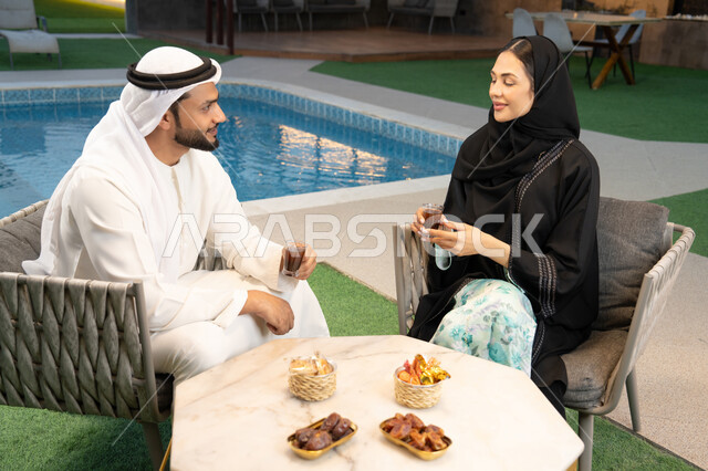 Drinking Arabic coffee, spending quality time with family, understanding and intellectual bonding between spouses, eating snacks and entertainment, sitting in one of the upscale Emirati chalets, an Arab Gulf Emirati man wearing a kandura and ghutra exchanging conversations with his wife in the open air