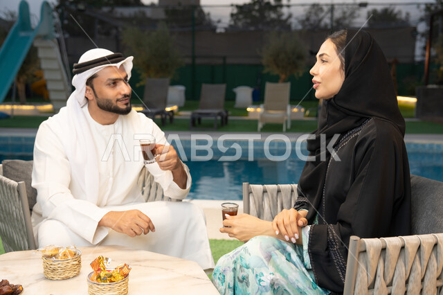 الجلوس في إحدى الشاليهات الاماراتية الراقية، رجل عربي خليجي اماراتي يرتدي الكندورة والغترة يقوم بتبادل الأحاديث مع زوجته في الهواء الطلق، شرب القهوة العربية، قضاء أوقات عائلية ممتعة، التفاهم والترابط الفكري بين الزوجين، تناول الوجبات الخفيفة والتسالي