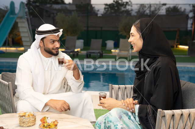 شرب القهوة العربية، قضاء أوقات عائلية ممتعة, التفاهم والترابط الفكري بين الزوجين، تناول الوجبات الخفيفة والتسالي، الجلوس في إحدى الشاليهات الاماراتية الراقية، رجل عربي خليجي اماراتي يرتدي الكندورة والغترة يقوم بتبادل الأحاديث مع زوجته في الهواء الطلق