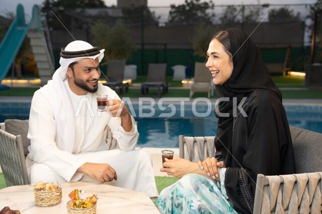 Understanding and intellectual connection between spouses, eating snacks and entertainment, sitting in one of the upscale Emirati chalets, an Arab Gulf Emirati man wearing a kandura and ghutra exchanging conversations with his wife in the open air, drinking Arabic coffee, spending enjoyable family time.
