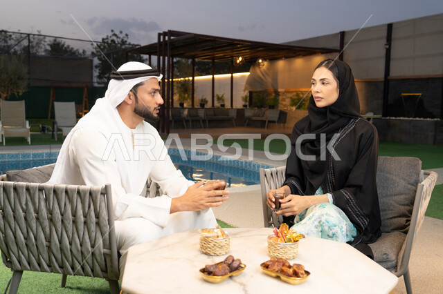 شرب القهوة العربية، قضاء أوقات عائلية ممتعة, التفاهم والترابط الفكري بين الزوجين، تناول الوجبات الخفيفة والتسالي، الجلوس في إحدى الشاليهات الاماراتية الراقية، رجل عربي خليجي اماراتي يرتدي الكندورة والغترة يقوم بتبادل الأحاديث مع زوجته في الهواء الطلق