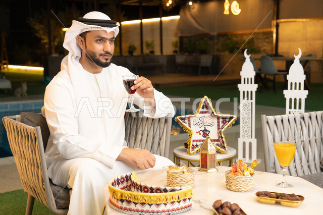 الاستمتاع بتناول العصائر الباردة، زينة واضاءة رمضانية، الجلسة الخارجية في احدى شاليهات الامارات، ضيافة شهر رمضان المبارك، شاب عربي خليجي اماراتي يرتدي الكندورة والغترة يجلس في الهواء الطلق يقم بشرب عصير التوت بتعابير السرور