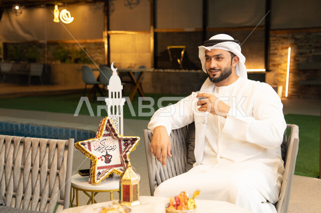 الاستمتاع بتناول العصائر الباردة، زينة واضاءة رمضانية، الجلسة الخارجية في احدى شاليهات الامارات، ضيافة شهر رمضان المبارك، شاب عربي خليجي اماراتي يرتدي الكندورة والغترة يجلس في الهواء الطلق يقم بشرب عصير التوت بتعابير السرور