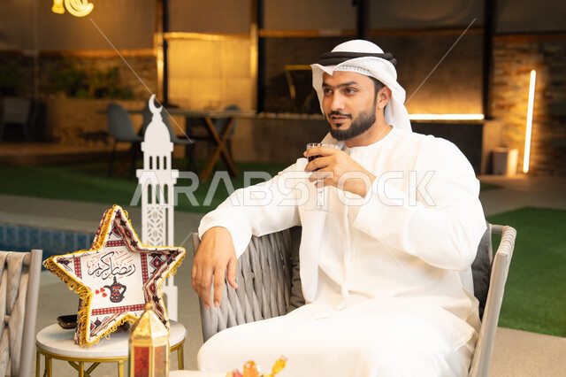 Enjoying cold juices, Ramadan decorations and lighting, outdoor seating in one of the chalets in the Emirates, hospitality of the holy month of Ramadan, a young Emirati Gulf Arab man wearing a kandura and ghutra sitting in the open air drinking berry juice with expressions of pleasure