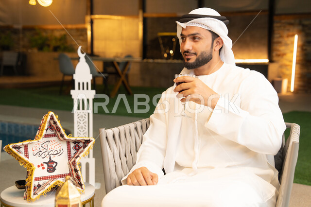 الاستمتاع بتناول العصائر الباردة، زينة واضاءة رمضانية، الجلسة الخارجية في احدى شاليهات الامارات، ضيافة شهر رمضان المبارك، شاب عربي خليجي اماراتي يرتدي الكندورة والغترة يجلس في الهواء الطلق يقم بشرب عصير التوت بتعابير السرور