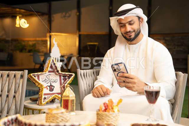 الجلسة الليلة الخارجية في احدى شاليهات الامارات، ضيافة شهر رمضان المبارك، شاب عربي خليجي اماراتي يرتدي الكندورة والغترة يجلس في الهواء الطلق، استخدام الهاتف المحمول بتعابير الاندماج والسرور، زينة واضاءة رمضانية