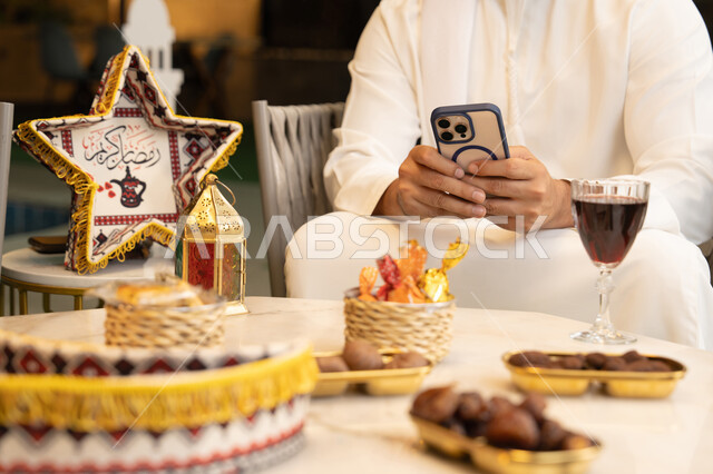 الجلسة الليلة الخارجية في احدى شاليهات الامارات، ضيافة شهر رمضان المبارك، شاب عربي خليجي اماراتي يرتدي الكندورة والغترة يجلس في الهواء الطلق، استخدام الهاتف المحمول بتعابير الاندماج والسرور، زينة واضاءة رمضانية
