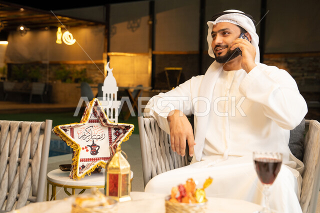 استخدام جهاز تقني حديث، شاب عربي خليجي اماراتي يرتدي الكندورة والغترة يجري مكالمة هاتفية عبر الهاتف المحمول يجلس في الهواء الطلق، زينة واضاءة رمضانية، الجلسة الليلة الخارجية في احدى الشاليهات، الرد على الاتصال الهاتفي بتعابير السرور، ضيافة شهر رمضان المبارك