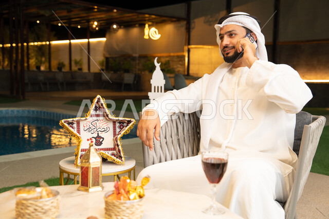 استخدام جهاز تقني حديث، شاب عربي خليجي اماراتي يرتدي الكندورة والغترة يجري مكالمة هاتفية عبر الهاتف المحمول يجلس في الهواء الطلق، زينة واضاءة رمضانية، الجلسة الليلة الخارجية في احدى الشاليهات، الرد على الاتصال الهاتفي بتعابير السرور، ضيافة شهر رمضان المبارك