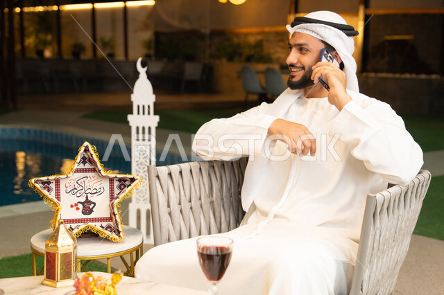 استخدام جهاز تقني حديث، شاب عربي خليجي اماراتي يرتدي الكندورة والغترة يجري مكالمة هاتفية عبر الهاتف المحمول يجلس في الهواء الطلق، زينة واضاءة رمضانية، الجلسة الليلة الخارجية في احدى الشاليهات، الرد على الاتصال الهاتفي بتعابير السرور، ضيافة شهر رمضان المبارك