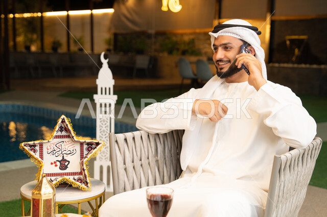 Using a modern technical device, a young Emirati Gulf Arab man wearing a kandura and ghutra making a phone call on his mobile phone sitting outdoors, Ramadan decorations and lighting, sitting outdoors at night in a chalet, answering the phone call with expressions of happiness, hospitality during the holy month of Ramadan