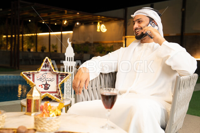 استخدام جهاز تقني حديث، شاب عربي خليجي اماراتي يرتدي الكندورة والغترة يجري مكالمة هاتفية عبر الهاتف المحمول يجلس في الهواء الطلق، زينة واضاءة رمضانية، الجلسة الليلة الخارجية في احدى الشاليهات، الرد على الاتصال الهاتفي بتعابير السرور، ضيافة شهر رمضان المبارك