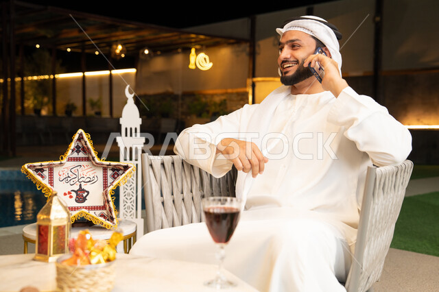 استخدام جهاز تقني حديث، شاب عربي خليجي اماراتي يرتدي الكندورة والغترة يجري مكالمة هاتفية عبر الهاتف المحمول يجلس في الهواء الطلق، زينة واضاءة رمضانية، الجلسة الليلة الخارجية في احدى الشاليهات، الرد على الاتصال الهاتفي بتعابير السرور، ضيافة شهر رمضان المبارك