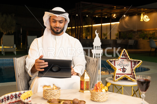 العمل عن بعد، الجلسة الليلة الخارجية في احدى شاليهات الامارات، ضيافة شهر رمضان المبارك، شاب عربي خليجي اماراتي يرتدي الكندورة والغترة يجلس في الهواء الطلق يعمل على جهاز لوحي، استخدام جهاز التابلت بتعابير الاندماج والسرور، زينة واضاءة رمضانية
