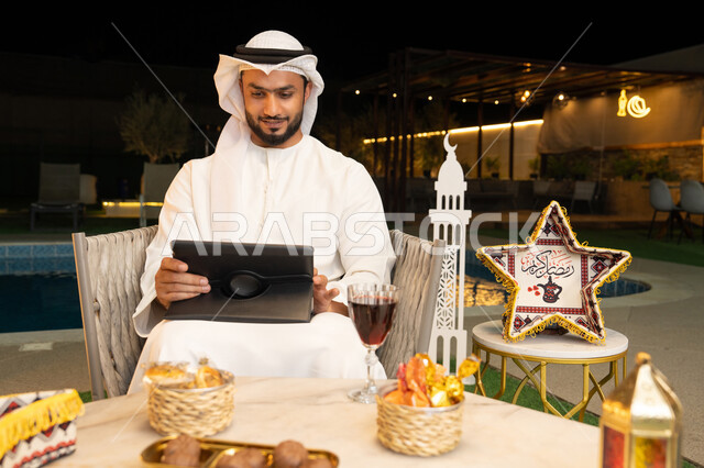 زينة واضاءة رمضانية، العمل عن بعد، الجلسة الليلة الخارجية في احدى شاليهات الامارات، ضيافة شهر رمضان المبارك، شاب عربي خليجي اماراتي يرتدي الكندورة والغترة يجلس في الهواء الطلق يعمل على جهاز لوحي، استخدام جهاز التابلت بتعابير الاندماج والسرور