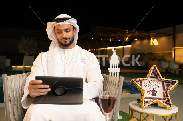 العمل عن بعد، الجلسة الليلة الخارجية في احدى شاليهات الامارات، ضيافة شهر رمضان المبارك، شاب عربي خليجي اماراتي يرتدي الكندورة والغترة يجلس في الهواء الطلق يعمل على جهاز لوحي، استخدام جهاز التابلت بتعابير الاندماج والسرور، زينة واضاءة رمضانية