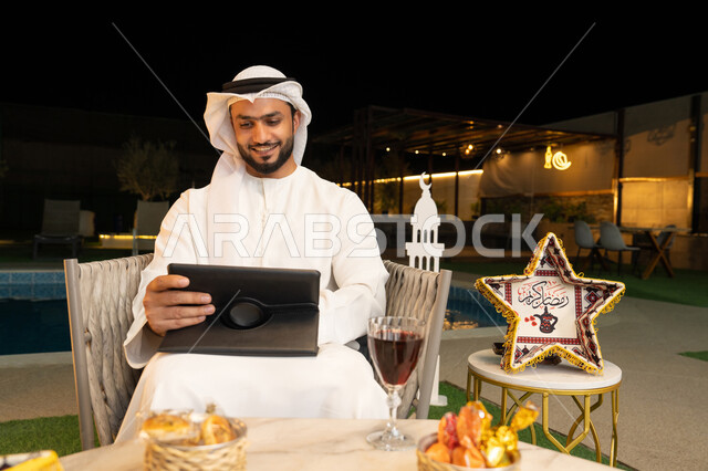 زينة واضاءة رمضانية، العمل عن بعد، الجلسة الليلة الخارجية في احدى شاليهات الامارات، ضيافة شهر رمضان المبارك، شاب عربي خليجي اماراتي يرتدي الكندورة والغترة يجلس في الهواء الطلق يعمل على جهاز لوحي، استخدام جهاز التابلت بتعابير الاندماج والسرور