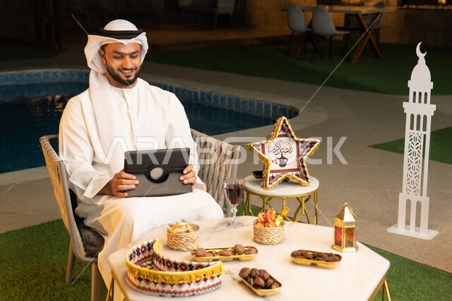 العمل عن بعد، الجلسة الليلة الخارجية في احدى شاليهات الامارات، ضيافة شهر رمضان المبارك، شاب عربي خليجي اماراتي يرتدي الكندورة والغترة يجلس في الهواء الطلق يعمل على جهاز لوحي، استخدام جهاز التابلت بتعابير الاندماج والسرور، زينة واضاءة رمضانية