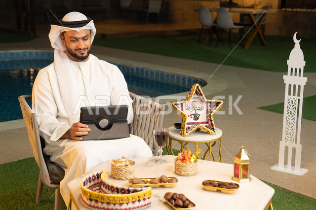 زينة واضاءة رمضانية، العمل عن بعد، الجلسة الليلة الخارجية في احدى شاليهات الامارات، ضيافة شهر رمضان المبارك، شاب عربي خليجي اماراتي يرتدي الكندورة والغترة يجلس في الهواء الطلق يعمل على جهاز لوحي، استخدام جهاز التابلت بتعابير الاندماج والسرور