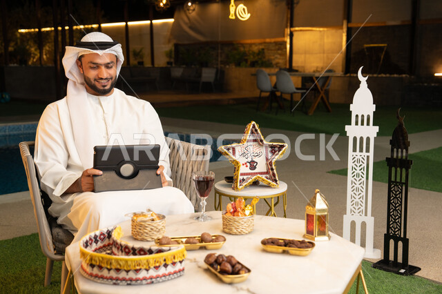 زينة واضاءة رمضانية، العمل عن بعد، الجلسة الليلة الخارجية في احدى شاليهات الامارات، ضيافة شهر رمضان المبارك، شاب عربي خليجي اماراتي يرتدي الكندورة والغترة يجلس في الهواء الطلق يعمل على جهاز لوحي، استخدام جهاز التابلت بتعابير الاندماج والسرور