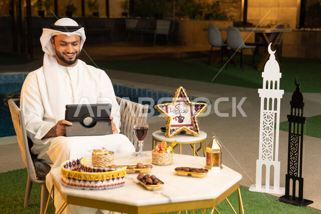 العمل عن بعد، الجلسة الليلة الخارجية في احدى شاليهات الامارات، ضيافة شهر رمضان المبارك، شاب عربي خليجي اماراتي يرتدي الكندورة والغترة يجلس في الهواء الطلق يعمل على جهاز لوحي، استخدام جهاز التابلت بتعابير الاندماج والسرور، زينة واضاءة رمضانية